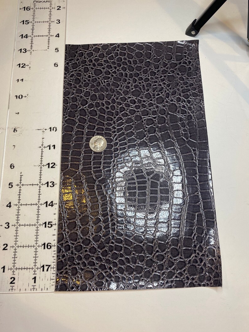 K&ouml;nnte beinhalten: Nahaufnahme eines dunkelgrauen Kunstkrokodillederstoffs. Der Stoff hat eine gl&auml;nzende, fast metallische Oberfl&auml;che. Der Stoff wird mit einem Lineal in Zoll gemessen.