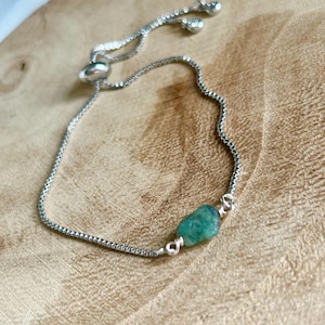 May include: Silver-toned adjustable bracelet featuring a small, raw emerald gemstone. The bracelet has a delicate chain design and a sliding bead closure. The emerald is a vibrant green color, adding a pop of color to the piece. The bracelet is displayed on a wooden surface.