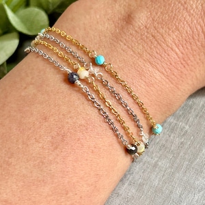 May include: Four delicate chain bracelets in gold and silver tones, each adorned with small, colorful beads. The bracelets feature turquoise, white, and dark blue beads. The bracelets are layered on a wrist, showcasing the jewelry's design.