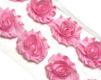 May include: Pink fabric rose trim on white mesh. The roses are a soft pink color and are evenly spaced along the trim. The trim is ideal for embellishing clothing, accessories, or craft projects.