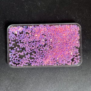 May include: A clear, rectangular container filled with small, star-shaped glitter in shades of pink and purple. The glitter is densely packed, creating a shimmering effect. The container has rounded corners.