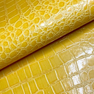 May include: Close-up of a yellow faux leather fabric with a crocodile embossed pattern. The fabric has a glossy finish.