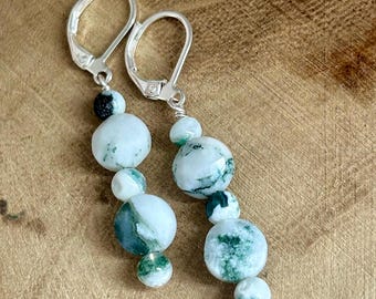 May include: A pair of dangle earrings featuring a series of round, white and green beads. The beads are strung together with silver-colored metal wire and attached to silver lever-back ear wires. The earrings have a natural, organic aesthetic.