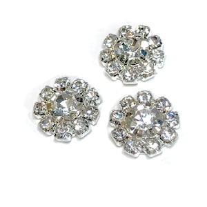 May include: Three silver-toned, rhinestone-encrusted flower-shaped embellishments. Each embellishment features a central clear rhinestone surrounded by smaller rhinestones.