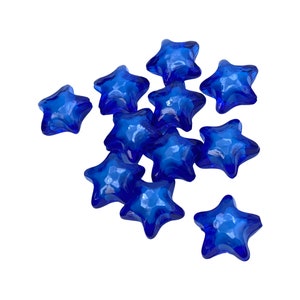 May include: A collection of eleven faceted, star-shaped glass beads in a deep, vibrant blue. The beads are scattered across a white background, each catching the light and displaying a textured surface. These decorative beads are ideal for crafting and jewelry making.