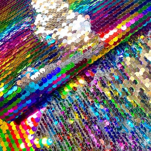 May include: Close-up of a fabric covered in iridescent sequins. The sequins are arranged in rows and change color from silver to a rainbow of hues. A section of the fabric has been flipped to reveal a silver color.