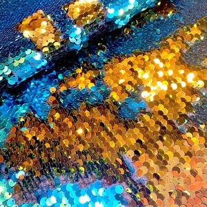 May include: Close-up of a fabric covered in iridescent sequins. The sequins are in shades of blue, gold, and teal, creating a shimmering effect. The fabric appears to be folded, showing the texture and depth of the material.