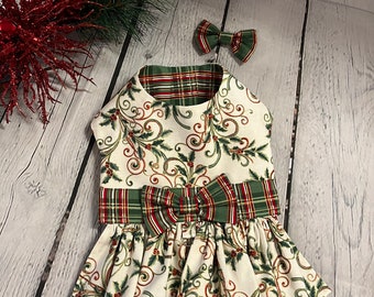 Puede incluir: Un vestido festivo para perros con pajarita a juego. El vestido presenta un fondo blanco con un diseño de acebo y bayas, acentuado por un lazo y ribete a cuadros verdes y rojos. Diseñado para un perro pequeño.