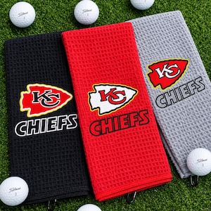 May include: Three rectangular waffle-weave golf towels in black, red, and gray, each featuring the Kansas City Chiefs logo and text. The towels are arranged with white Titleist golf balls scattered around them on a green surface.