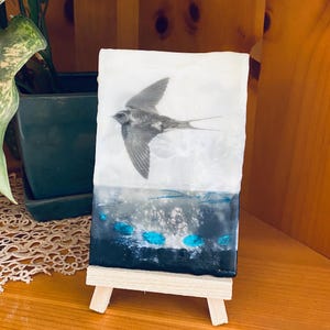 Op de afbeelding: Een klein, rechthoekig kunstwerk op een houten ezel. Het kunstwerk toont een grijswaardenafbeelding van een vogel in vlucht boven een donkerblauw en zwart abstract ontwerp met turquoise accenten. Gepresenteerd op een kleine houten ezel.