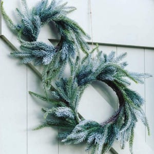 May include: Two artificial evergreen wreaths, dusted with white, hang against a pale green door. The wreaths are circular, with a natural, rustic look. Ideal for seasonal decoration, they offer a touch of festive charm.