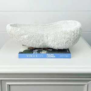 May include: A white, textured, coral-shaped decorative bowl. The bowl sits on a book titled "Vogue On Ralph Lauren". The bowl has a unique, organic shape and is made of a porous material. The background is a white wall and a white table.