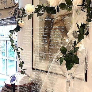 May include: A clear acrylic seating chart sign with white printed text that reads Please find your seat, followed by lists of names organised by letter. The rectangular sign is decorated with artificial white roses and green leaves.