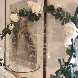May include: A transparent acrylic wedding sign with white cursive lettering. The sign says "Welcome to Harriet & Nicholas Unplugged Ceremony." It is adorned with white roses and green foliage.