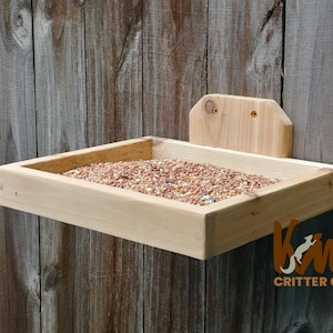 Puede incluir: Un comedero para pájaros de madera con una bandeja rectangular llena de alpiste. El comedero está montado en una valla de madera. El comedero es de madera de color claro y tiene un pequeño respaldo. El logotipo "Kim's Critter Corner" es visible.