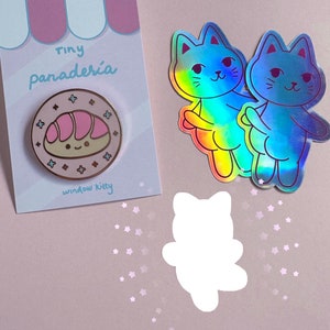 May include: A pink and white enamel pin featuring a cartoon bread roll with a happy face. The pin is surrounded by a blue circle with white stars. The pin is on a pink and white card with the text "tiny panadería" and "window kitty".