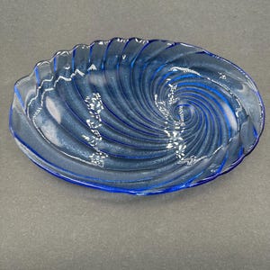 May include: A blue glass oval-shaped dish with a spiral design. The dish has a wavy edge and a deep blue color. The spiral pattern starts from the center and expands outwards. The dish is made of glass.