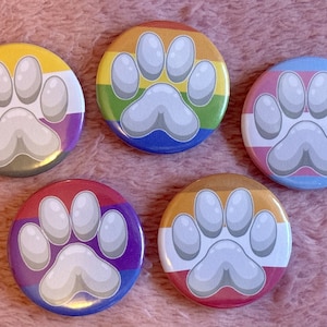 May include: Five circular badges with a paw print design, each representing a different LGBTQ+ pride flag: non-binary, rainbow, transgender, bisexual, and lesbian. The badges are displayed on a pink, fluffy surface.