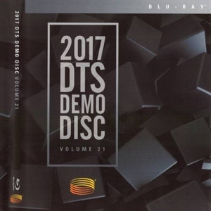 Puede incluir: Un disco Blu-ray con el texto "2017 DTS DEMO DISC VOLUME 21" en blanco. El disco tiene un fondo negro con un patrón geométrico de cubos grises oscuros. El logotipo de DTS es visible en la esquina inferior izquierda.