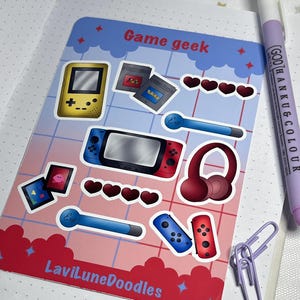 Op de afbeelding: Een stickervel met een "Game geek" thema, met illustraties van retro videogameconsoles, cartridges en controllers. Het vel heeft een rood en blauw kleurenschema, met de tekst "LaviLuneDoodles" onderaan. Een paarse pen en paperclips zijn ook zichtbaar.