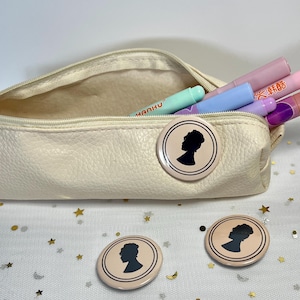Op de afbeelding: Een crèmekleurige etui met een rits, met kleurrijke lippenstiften en een mintgroene pen. Drie ronde, lichtroze knopen met een zwart silhouet van een vrouwenprofiel liggen op een witte ondergrond met gouden en zilveren sterrenconfetti.