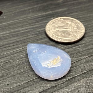 May include: A pale blue, teardrop-shaped gemstone next to a coin. The gemstone has a smooth, polished surface with light brown inclusions. The coin is silver. The gemstone is likely for jewelry making.