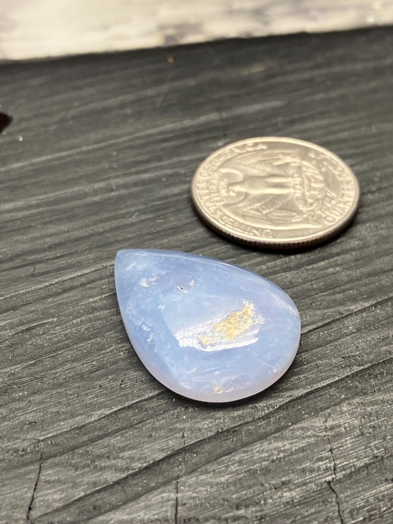 May include: A pale blue, teardrop-shaped gemstone next to a coin. The gemstone has a smooth, polished surface with light brown inclusions. The coin is silver. The gemstone is likely for jewelry making.