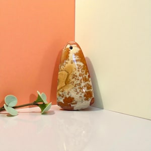May include: A polished, teardrop-shaped pendant with a hole at the top. The stone features a mix of tan, brown, and white patterns. The pendant is set against a peach and cream-colored backdrop.