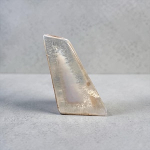 May include: A polished, triangular agate stone with a translucent, layered appearance. The stone features a gradient of colours, including white, beige, and hints of lavender. The edges are smooth, and the overall shape is geometric.