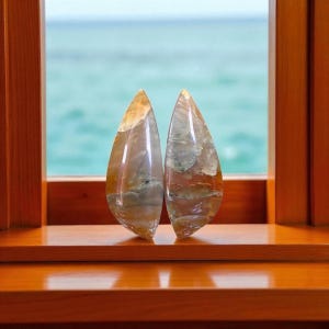 Peut inclure: Deux pierres précieuses polies en forme de goutte d'eau, aux couleurs variées, notamment beige, marron et vert. Les pierres sont exposées sur une surface en bois devant une fenêtre avec vue sur la mer.