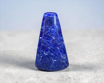 May include: A polished, deep blue sodalite gemstone, cone-shaped with a flat base. The stone has white veining throughout, creating a marbled effect. The background is a soft gray and blue.