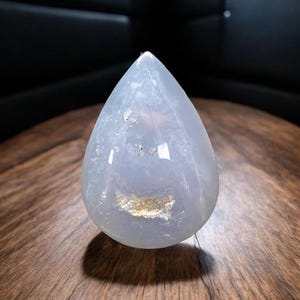 May include: A teardrop-shaped, translucent, pale blue gemstone with a polished surface. The stone has a light-coloured inclusion near the centre and is set against a dark background. The gemstone is displayed on a wooden surface.