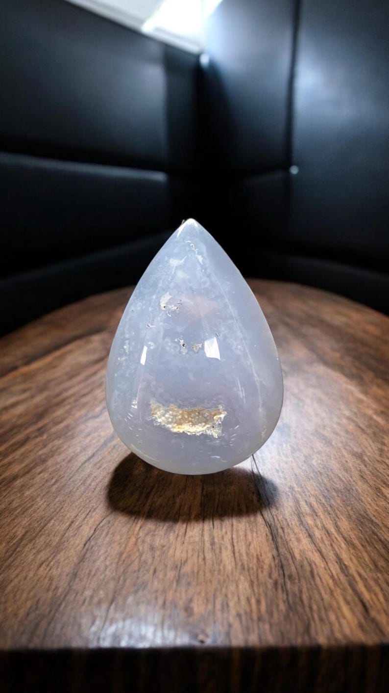 May include: A teardrop-shaped, translucent, pale blue gemstone with a polished surface. The stone has a light-coloured inclusion near the centre and is set against a dark background. The gemstone is displayed on a wooden surface.