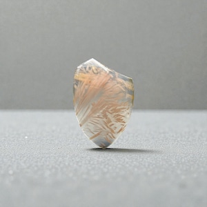 May include: A clear, shield-shaped gemstone with internal inclusions of feathery, golden-brown patterns. The stone is polished and translucent, with a slightly irregular top edge. The gemstone is set against a gray background.
