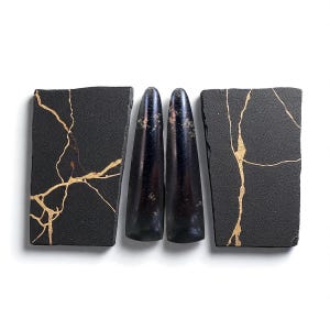 Puede incluir: Una composición de naturaleza muerta con dos rectángulos negros con vetas doradas, que enmarcan dos objetos oscuros, alargados y en forma de lágrima. Los objetos están dispuestos sobre una superficie blanca, creando un contraste llamativo.