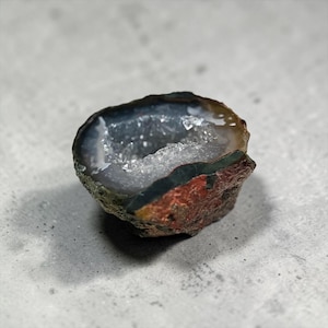 Peut inclure: Une géode coupée en deux, révélant un intérieur cristallin. L'extérieur présente un mélange de couleurs, notamment marron, rouge, vert et gris. L'intérieur scintille de minuscules cristaux, créant un contraste saisissant.