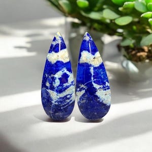 May include: Two teardrop-shaped lapis lazuli cabochons with white veining. The stones are a deep blue with white streaks and swirls.