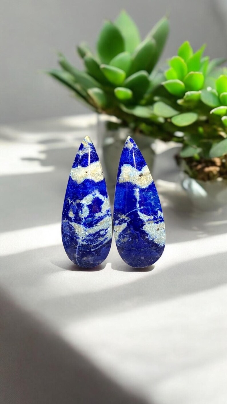 May include: Two teardrop-shaped lapis lazuli cabochons with white veining. The stones are a deep blue with white streaks and swirls.