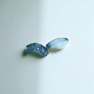 Puede incluir: Dos piedras de ágata azul pulidas de forma irregular. Las piedras tienen un borde exterior azul oscuro y un interior azul más claro, con unos pequeños agujeros. Las piedras están sobre un fondo blanco. Adecuado para joyería.