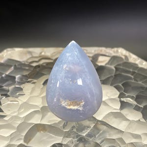 May include: A polished, teardrop-shaped gemstone in a pale blue colour. The stone has a smooth, glossy surface with subtle internal patterns and a small, beige inclusion. The gemstone is displayed against a textured, clear background.