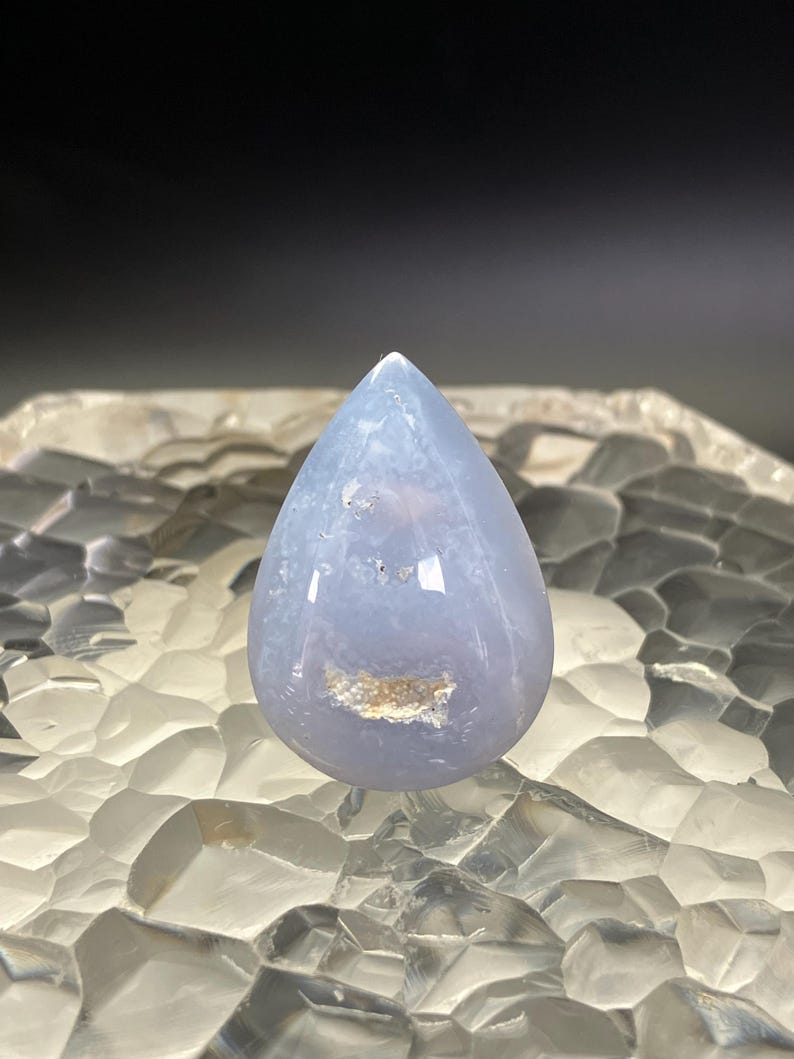 May include: A polished, teardrop-shaped gemstone in a pale blue colour. The stone has a smooth, glossy surface with subtle internal patterns and a small, beige inclusion. The gemstone is displayed against a textured, clear background.