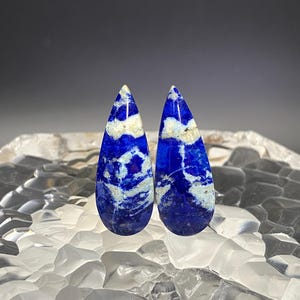 May include: Two teardrop-shaped lapis lazuli cabochons with white veins and a blue background.