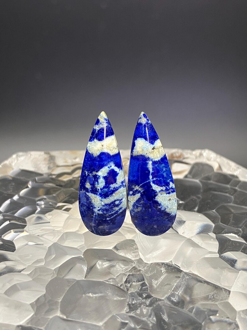 May include: Two teardrop-shaped lapis lazuli cabochons with white veins and a blue background.