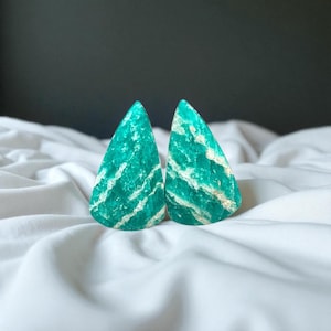 May include: Two teardrop-shaped Amazonite stones with a vibrant teal color and white veining. The stones are displayed on a white, textured fabric, showcasing their natural beauty and unique patterns. The gemstones are ideal for jewelry making.