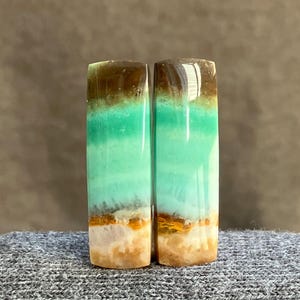 May include: Two polished gemstone slabs. Each slab is rectangular with rounded edges and features layers of brown, green, and tan colors. The stones are displayed on a gray surface.
