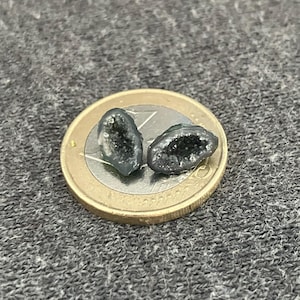 May include: Two small, dark grey geode halves with sparkling crystal interiors sit on a Euro coin. The geodes have a rough, textured exterior and a cavity filled with tiny, glittering crystals. The coin provides a size reference.