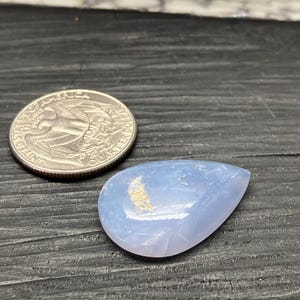 May include: A pale blue, teardrop-shaped gemstone with a polished surface. A U.S. quarter coin is next to the gemstone for size comparison. The gemstone has a light-coloured, crescent-shaped inclusion.