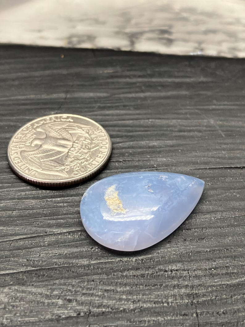 May include: A pale blue, teardrop-shaped gemstone with a polished surface. A U.S. quarter coin is next to the gemstone for size comparison. The gemstone has a light-coloured, crescent-shaped inclusion.