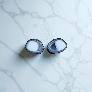 Puede incluir: Dos geodas de ágata pulidas, de forma ovalada, con interior blanco y gris y borde gris oscuro. Las geodas se exhiben sobre una superficie de mármol blanco. Las geodas son un producto de piedra natural.