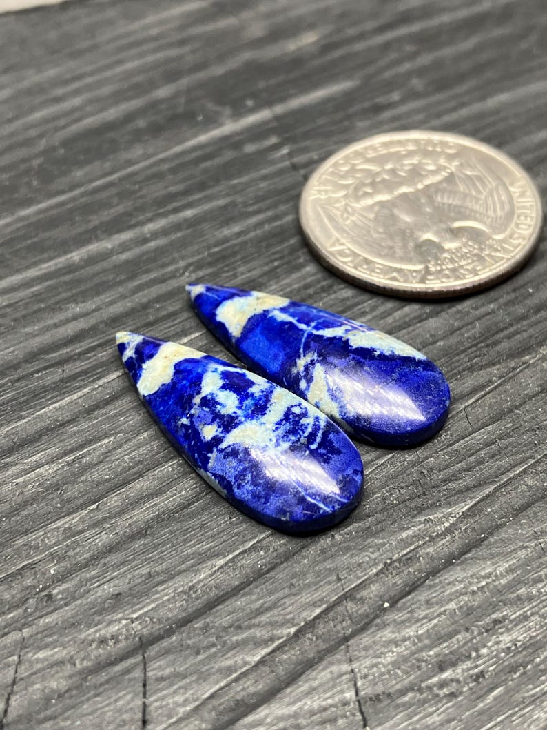May include: Two teardrop-shaped lapis lazuli cabochons with white veining. The stones are approximately the size of a US quarter.